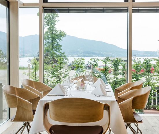 Elegant eingedeckte Tafel mit weißer Tischwaesche, Servietten, Glaeser und Besteck im Wintergarten des Stieler-Haus mit Blick durch die großen Fenster auf den Tegernsee