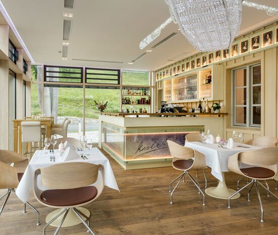 Wintergarten im Stieler-Haus Blick auf die Bar mit Tresen, Glaeser, Kaffeemaschine, Deckenlampe aus Kristall und an der Wand die gemalten Bilder der Damen der Schoenheitengalerie von Karl Stieler vorne eingedeckte Tische und Stuehle Boden aus Altholz