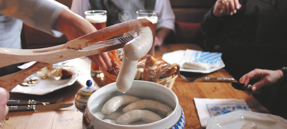 4 Stueck Weisswuerste in einer Schale mit warmen Wasser, zwei Stueck in einer Wurstzange aus Holz Korb mit Brezen und suesser Senf Menschen an einem Holztisch